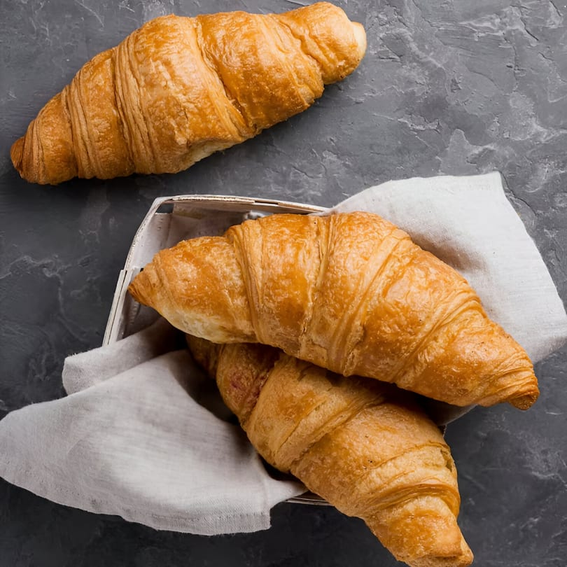 Golden-brown croissants, one on a dark gray surface and two in a white basket with a linen napkin.